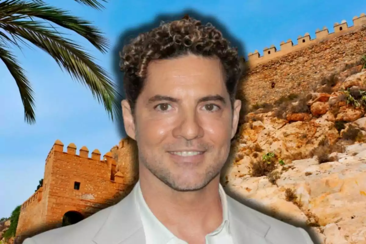 David Bisbal con cabello rizado y una chaqueta clara sonríe frente a un fondo de una fortaleza de piedra y una palmera bajo un cielo azul.