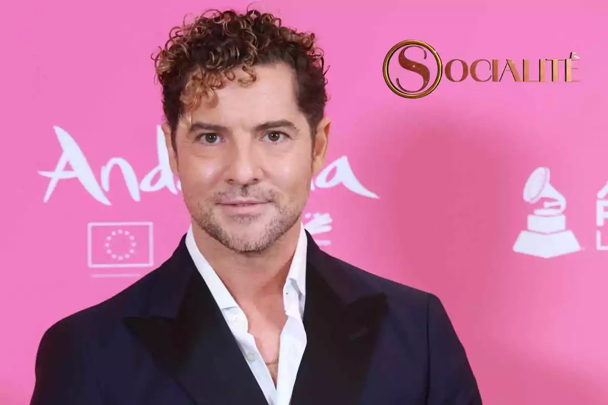 David Bisbal, vestido con un traje oscuro y camisa blanca, posando frente a un fondo rosa con logotipos y texto.