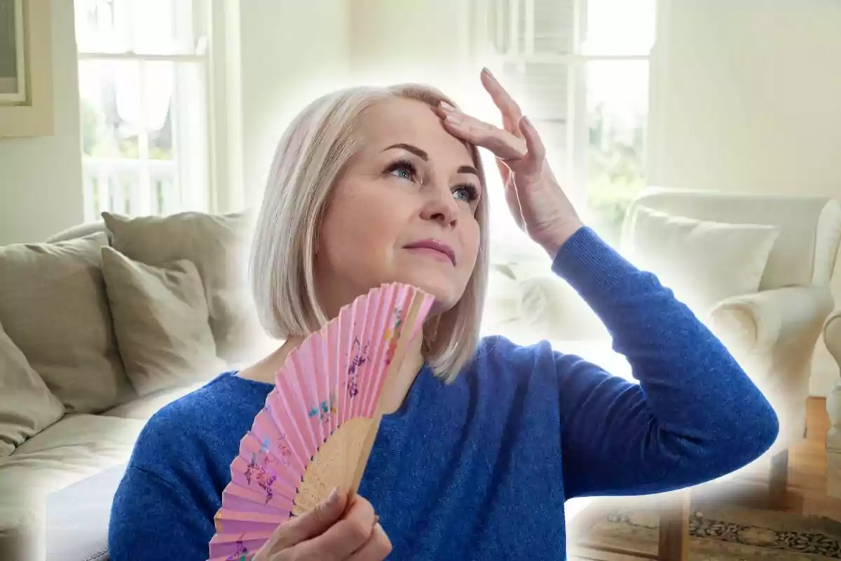 Mujer de cabello rubio corto usando un abanico rosa mientras se toca la frente en una sala de estar iluminada.