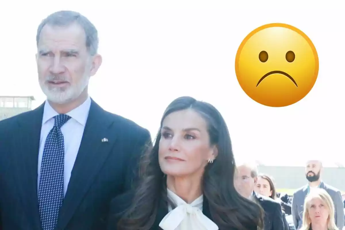 Felipe VI and Queen Letizia dressed formally are standing outdoors with a sad face emoji overlaid in the top right corner.