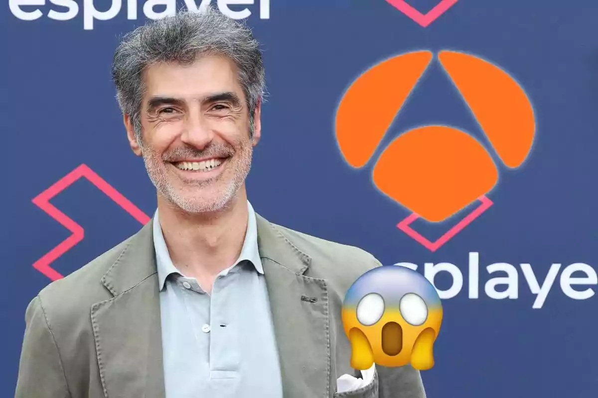 Hombre sonriente con cabello canoso y barba, vestido con una chaqueta verde y camisa gris, posando frente a un fondo azul con logotipos y un emoji de sorpresa en la esquina inferior derecha.