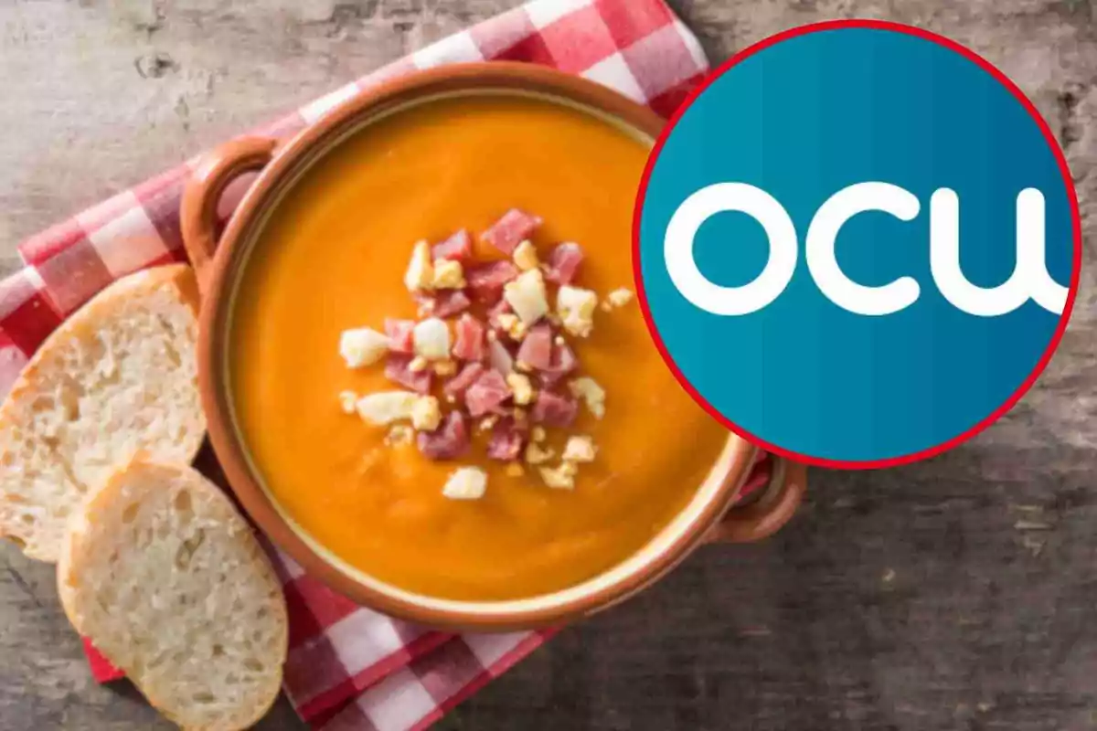 Un plato de sopa de color naranja con trozos de jamón y huevo en el centro acompañado de rebanadas de pan sobre un mantel a cuadros rojos y blancos con un logotipo circular azul y blanco en la esquina. Un plato de sopa de color naranja con trozos de jamón y huevo en el centro acompañado de rebanadas de pan sobre un mantel a cuadros rojos y blancos con un logotipo circular azul y blanco en la esquina.