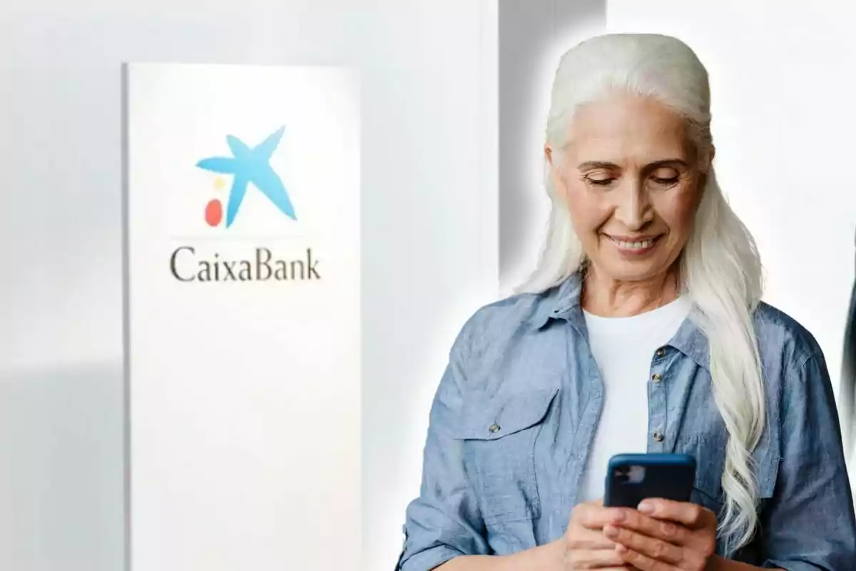 Una mujer mayor con cabello blanco sonríe mientras mira su teléfono móvil, con un cartel de CaixaBank al fondo.