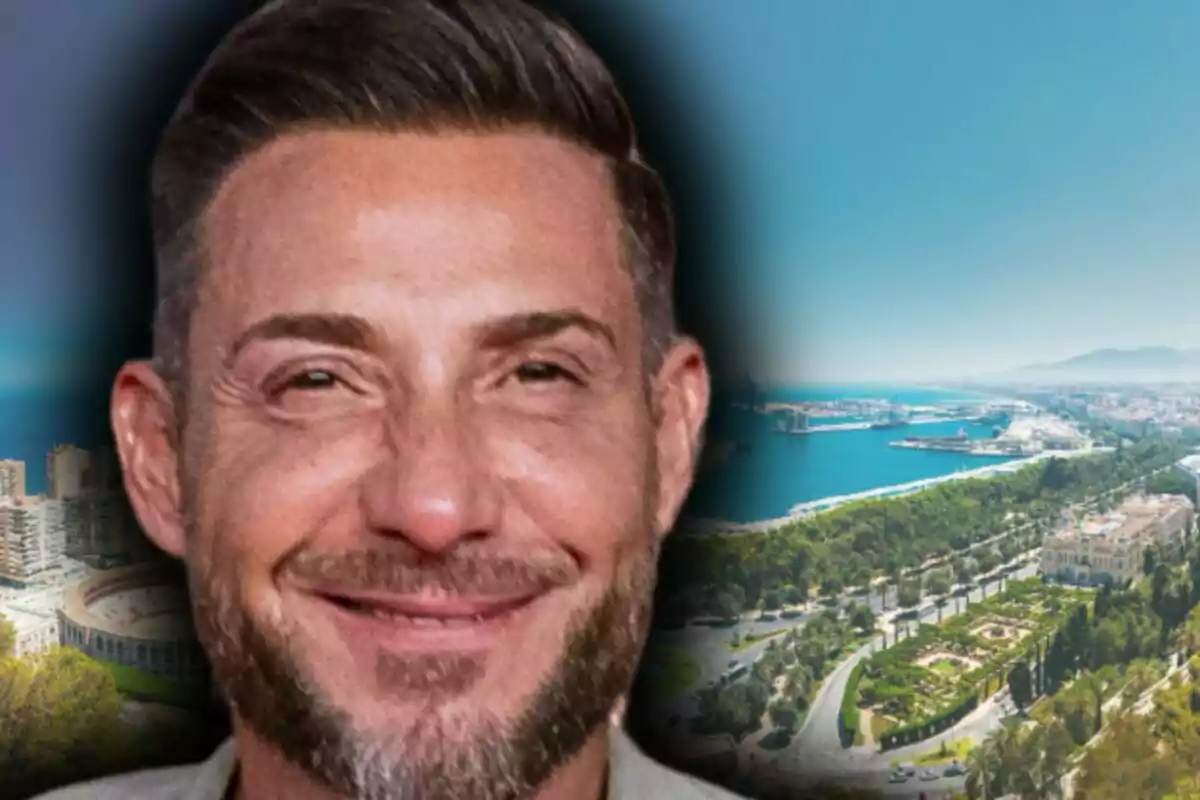Imagen de Antonio David Flores sonriente con barba y cabello corto que aparece en primer plano con un fondo de una ciudad costera y un puerto, la cual es Málaga.
