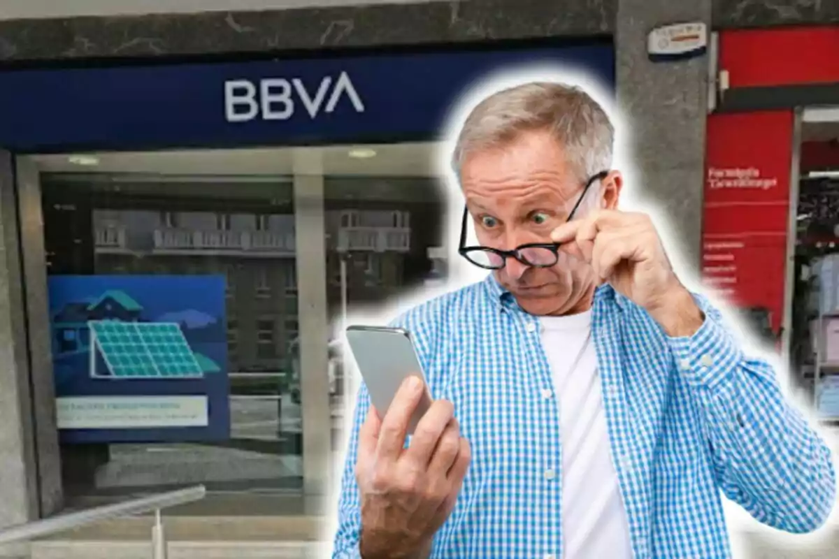 Un hombre mayor con camisa a cuadros azules y gafas observa sorprendido su teléfono móvil frente a una sucursal del banco BBVA.