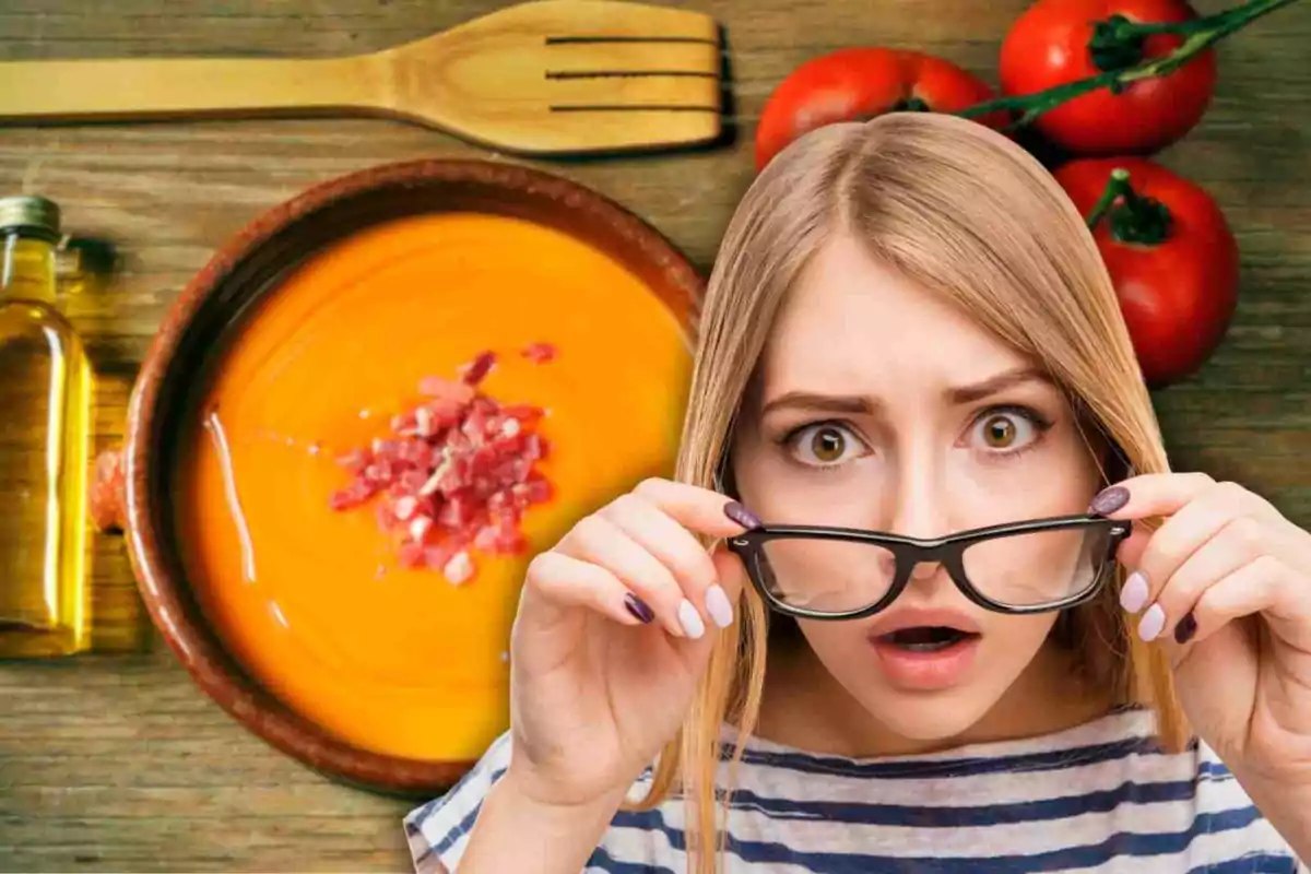 Una mujer con expresión de sorpresa sostiene sus gafas frente a un plato de sopa de tomate con trozos de jamón, acompañado de una botella de aceite, una cuchara de madera y tomates frescos sobre una mesa de madera.
