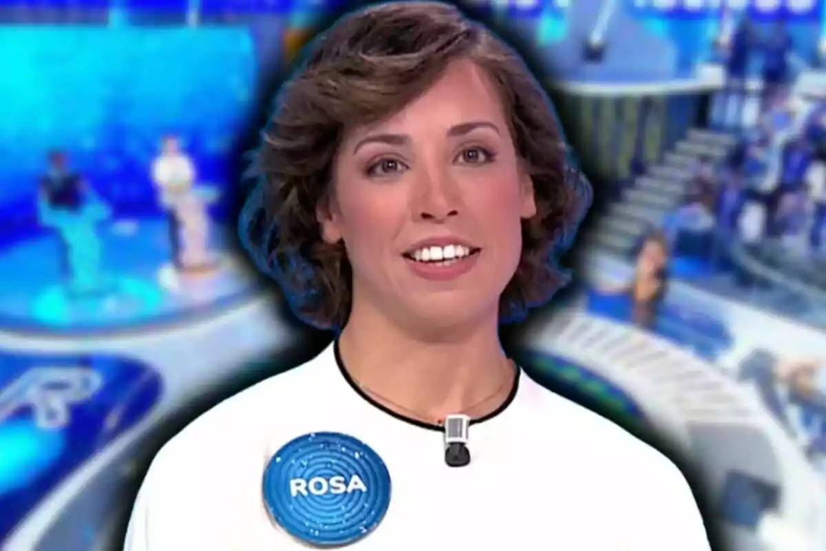 Rosa, concursante de 'Pasapalabra', sonriente con cabello corto y rizado lleva una camiseta blanca con un micrófono y una etiqueta que dice "Rosa" en un entorno de programa de televisión con un fondo azul desenfocado.