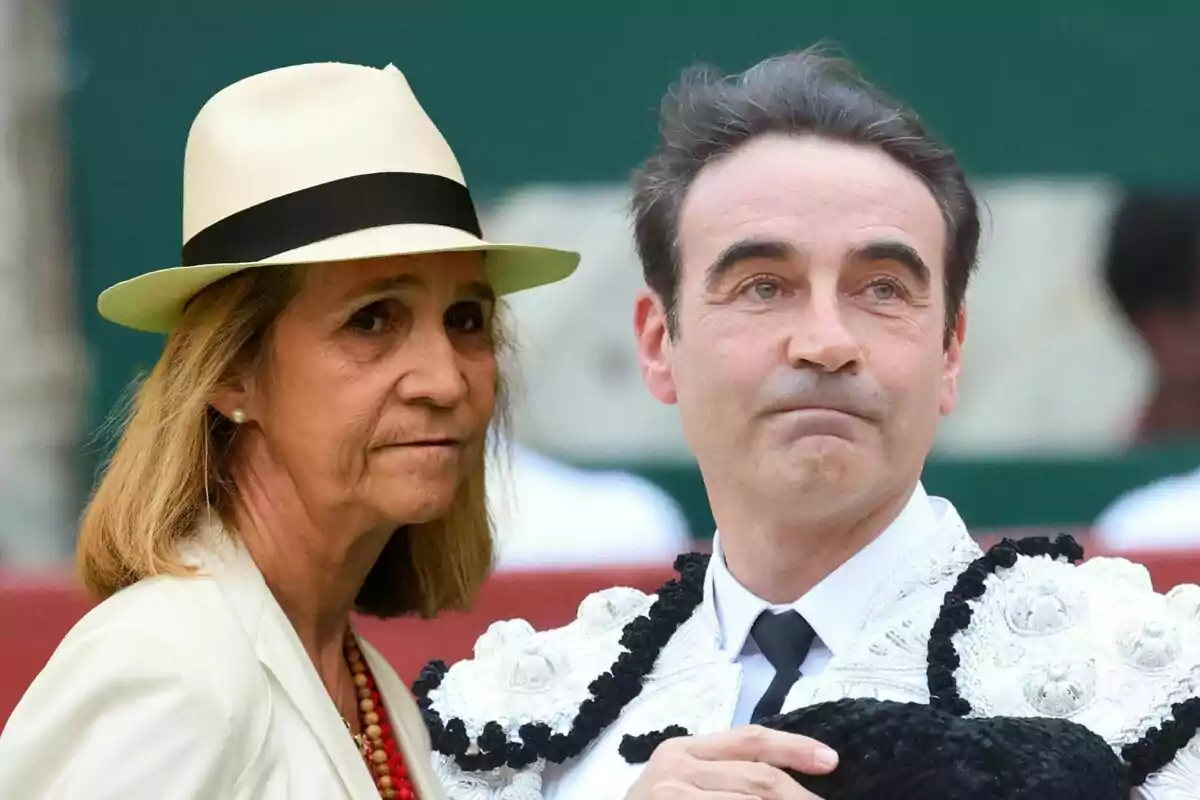 La infanta Elena con sombrero y Enrique Ponce vestido de torero están juntos en un entorno al aire libre.