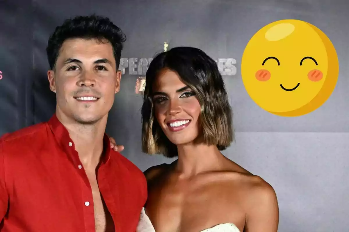 Kiko Jiménez and Sofía Suescun smiling while posing in front of a dark background with a yellow smiling emoji in the top right corner.