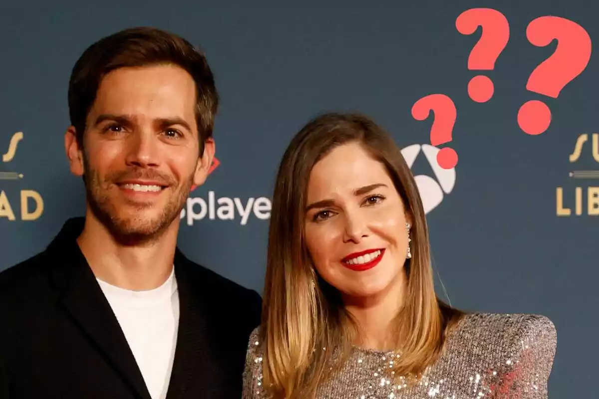 Marc Clotet y Natalia Sánchez sonriente posando en un evento con un fondo azul y signos de interrogación rojos sobre sus cabezas.