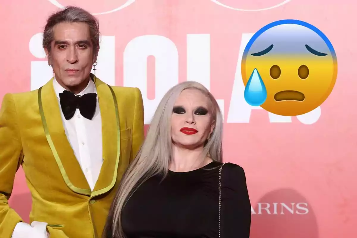 Mario Vaquerizo y Alaska posa en un evento con un fondo rojo y un emoji de cara preocupada.