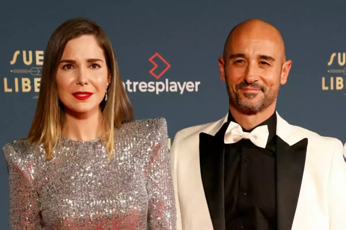 Natalia Sánchez con vestido plateado brillante y Alain Hernández con esmoquin blanco y negro posan juntos en un evento con fondo azul y logotipos.