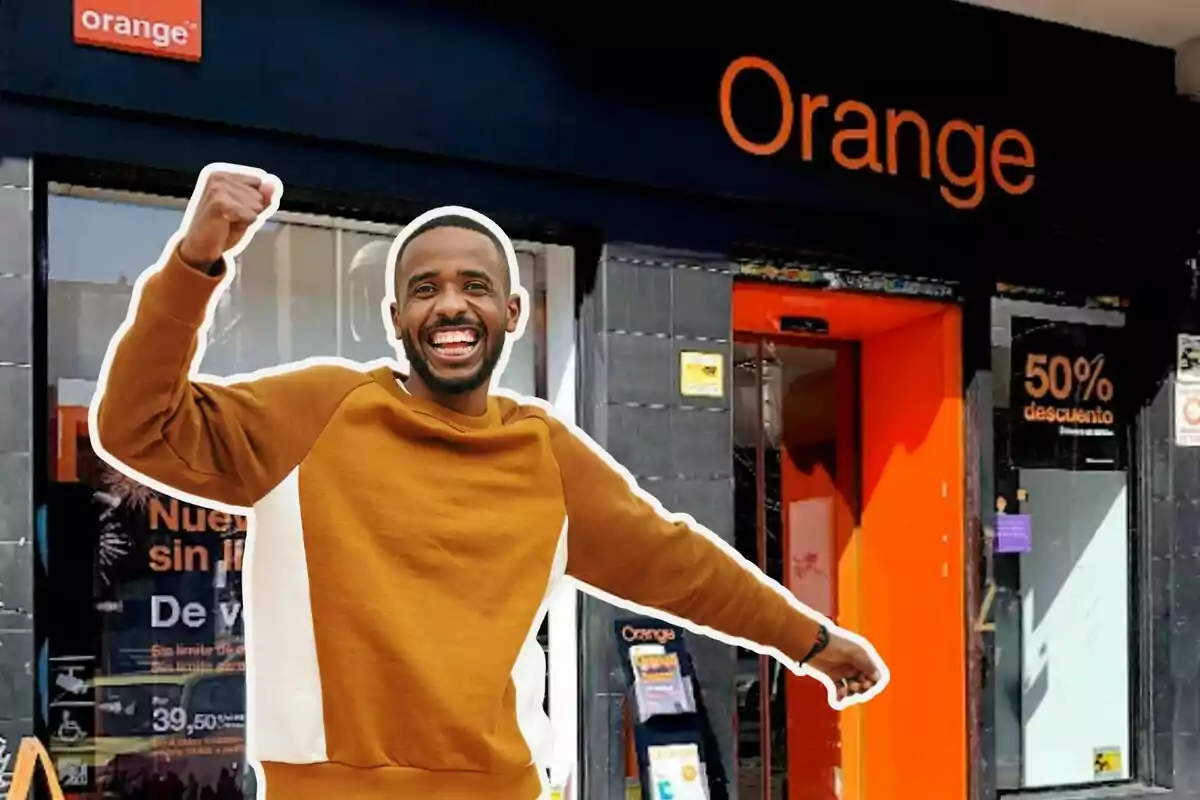 A smiling man in a brown sweater raises his fist in front of an Orange store.