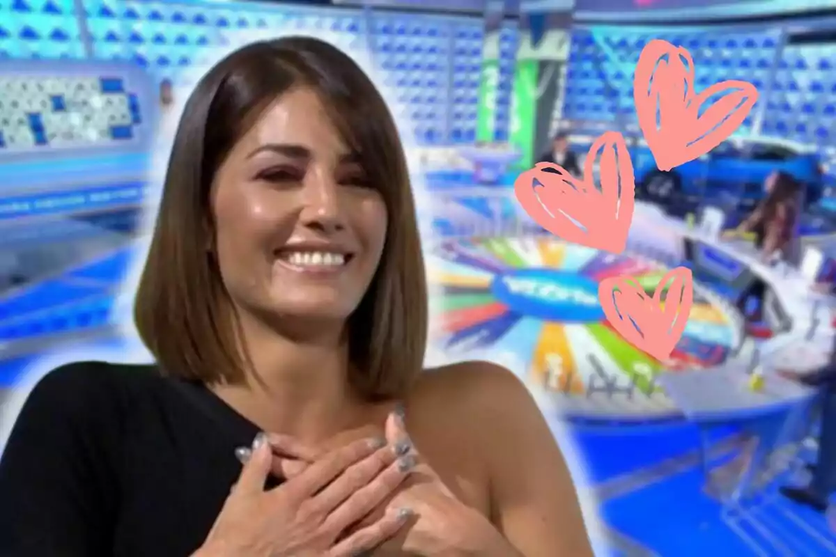 Laura Moure sonriente con las manos en el pecho en un set de televisión colorido con corazones dibujados.