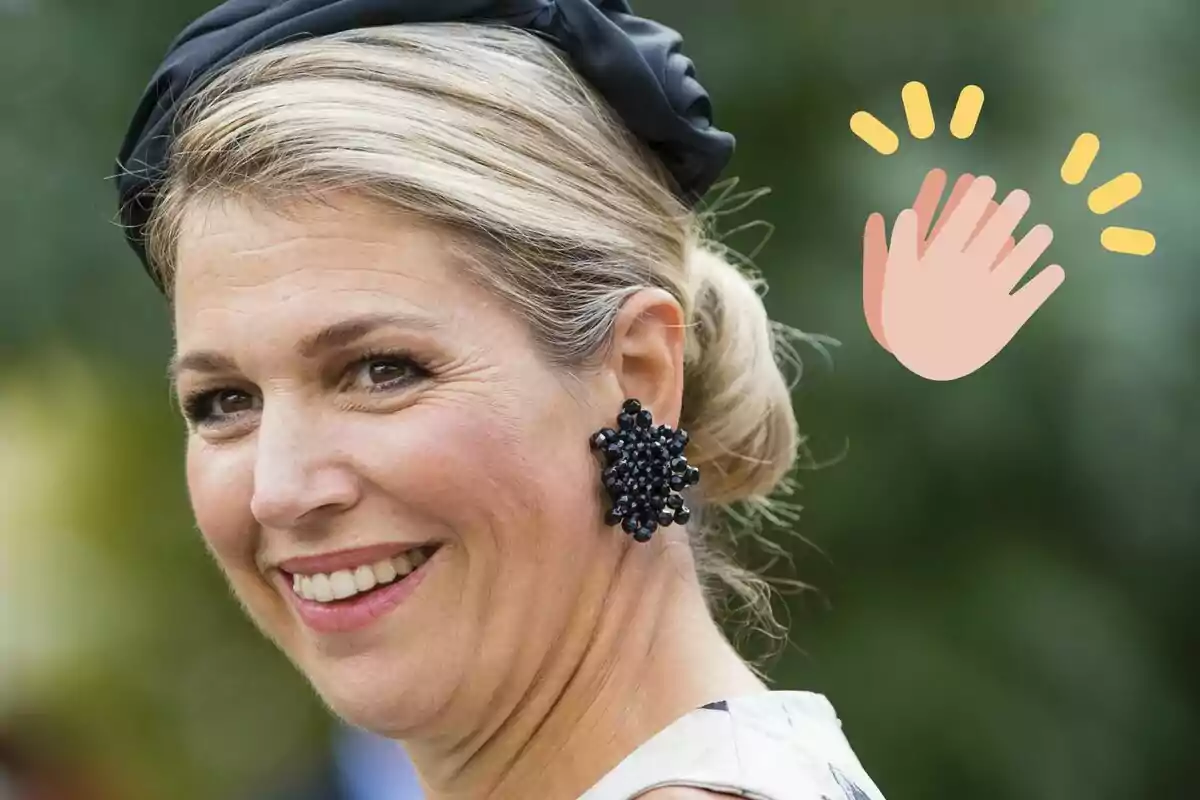 La reina Máxima de Holanda sonriente con un peinado recogido y un accesorio negro en el cabello lleva un pendiente grande y oscuro mientras un emoji de aplauso aparece al lado.