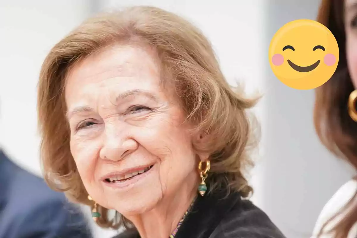 Queen Sofia smiling with light brown hair and gold earrings next to a smiley emoji.