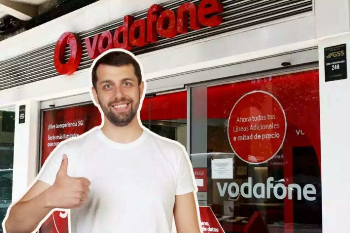 A smiling man in a white T-shirt and thumbs up stands in front of a Vodafone store.