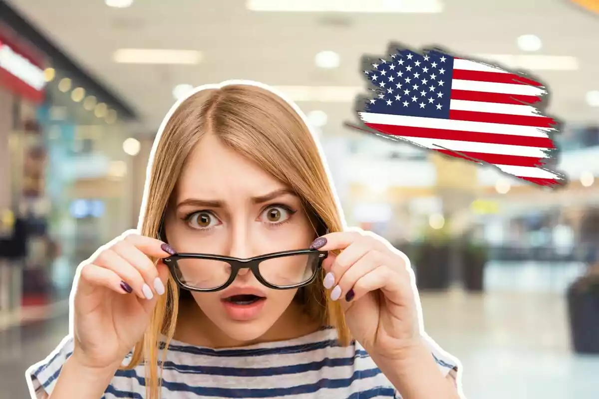 A woman with a surprised expression holds her glasses in an indoor setting with a United States flag in the background.