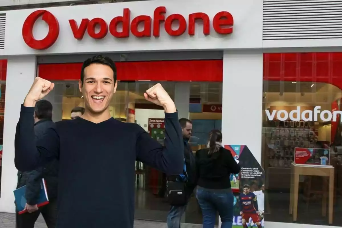 A smiling man with his arms raised in front of a Vodafone store.