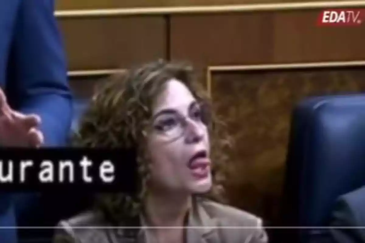 Una persona con gafas y cabello rizado está sentada en un entorno formal, mirando hacia arriba con la boca abierta, mientras un subtítulo parcialmente visible aparece en la parte inferior de la imagen. Una persona con gafas y cabello rizado está sentada en un entorno formal, mirando hacia arriba con la boca abierta, mientras un subtítulo parcialmente visible aparece en la parte inferior de la imagen.