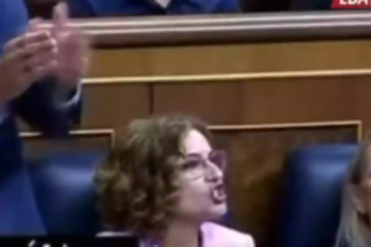 Mujer con gafas y cabello rizado hablando enérgicamente en un recinto parlamentario mientras una persona aplaude cerca de ella Mujer con gafas y cabello rizado hablando enérgicamente en un recinto parlamentario mientras una persona aplaude cerca de ella