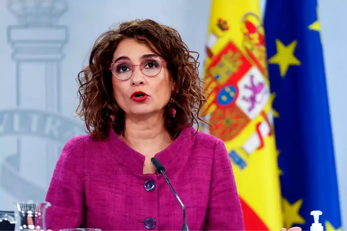 Una mujer con gafas y chaqueta morada hablando frente a un micrófono con las banderas de España y la Unión Europea de fondo.