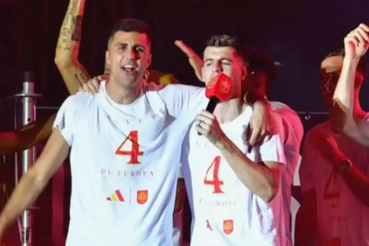 Dos personas con camisetas blancas con el número 4 celebran en un escenario, una de ellas sostiene un micrófono rojo.