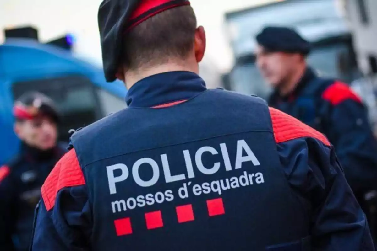 Policías de los Mossos d'Esquadra en uniforme, de espaldas, con vehículos policiales en el fondo.