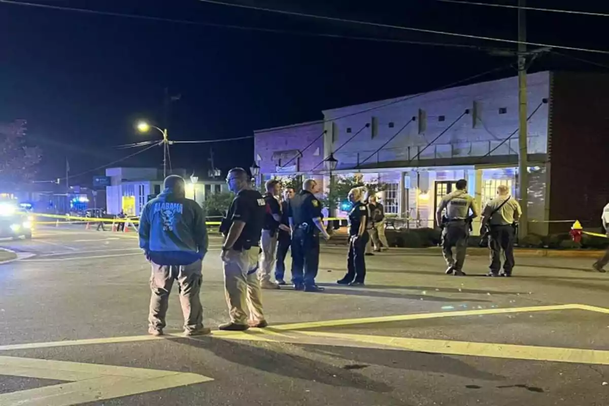 Varias personas, incluyendo oficiales de policía, están reunidas en una calle acordonada por la noche, con luces de emergencia y edificios al fondo.