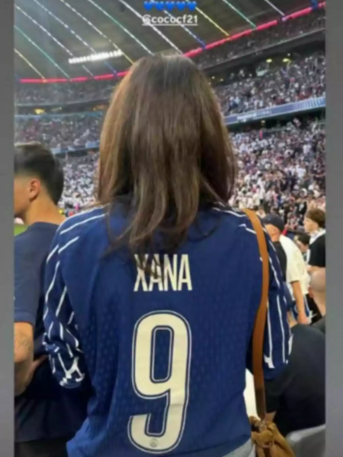 Mujer de espaldas con camiseta azul de fútbol que lleva el nombre Xana y el número 9 en un estadio lleno de espectadores Mujer de espaldas con camiseta azul de fútbol que lleva el nombre Xana y el número 9 en un estadio lleno de espectadores