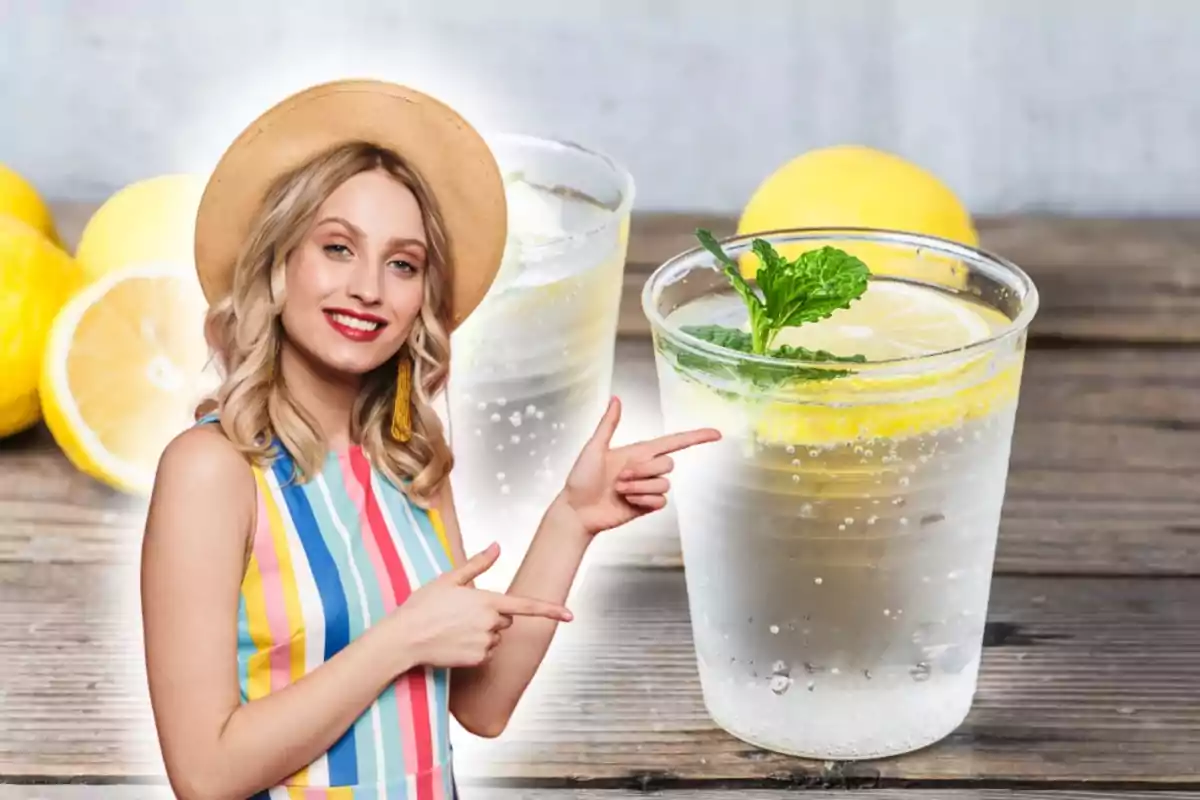 Una mujer con sombrero y vestido de rayas coloridas sonríe y señala hacia dos vasos de limonada con rodajas de limón y hojas de menta, con limones en el fondo sobre una mesa de madera.