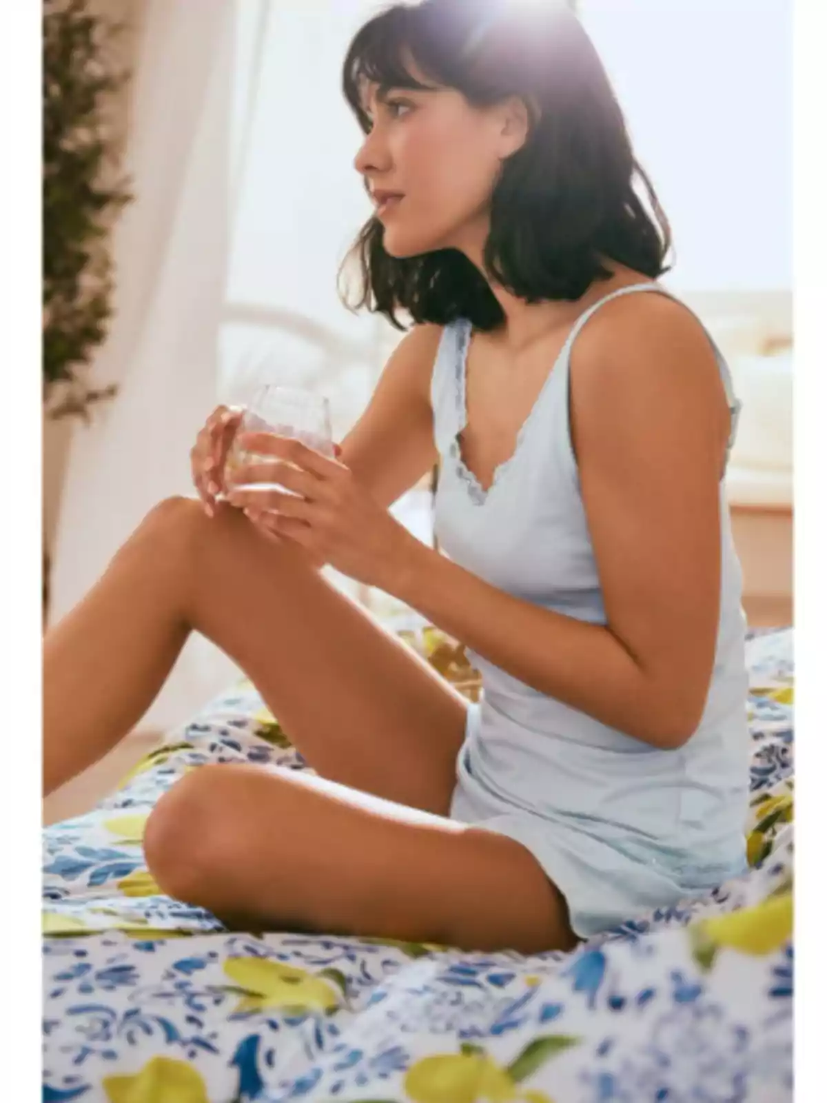 Mujer sentada en una cama con ropa de dormir clara sosteniendo un vaso y mirando hacia un lado en una habitación iluminada Mujer sentada en una cama con ropa de dormir clara sosteniendo un vaso y mirando hacia un lado en una habitación iluminada