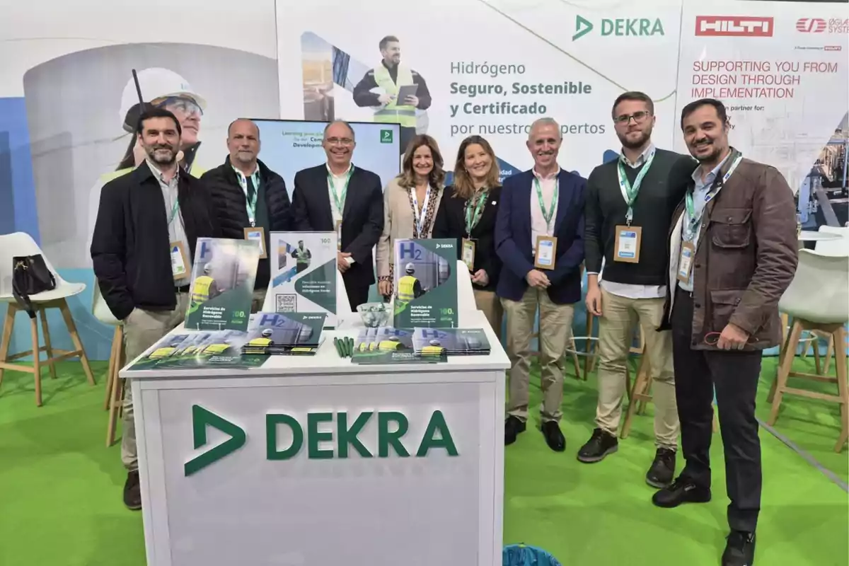 Un grupo de personas posando en un stand de exhibición de DEKRA con folletos y material promocional sobre hidrógeno seguro y sostenible.