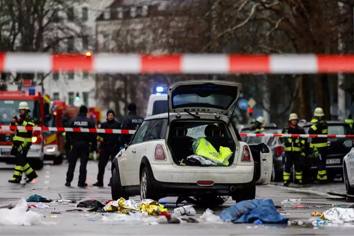 A car with the trunk open on a cordoned-off street, surrounded by police and firefighters, with debris on the ground.