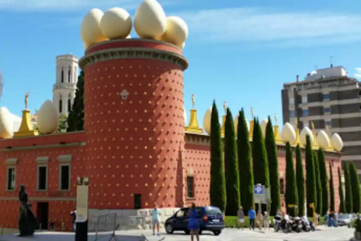 Edificio con fachada roja decorada con figuras doradas y coronado con grandes esculturas de huevos, rodeado de árboles y personas caminando.