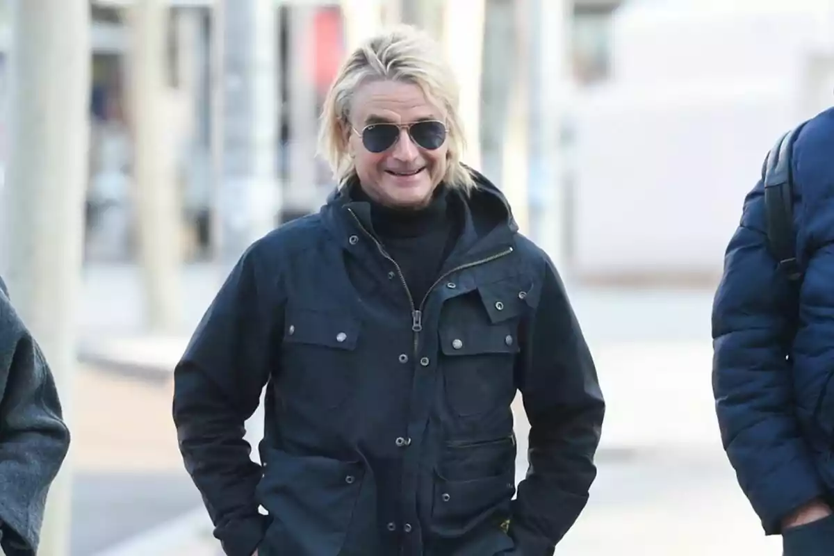 A man with sunglasses and a dark jacket smiles as he walks down the street.