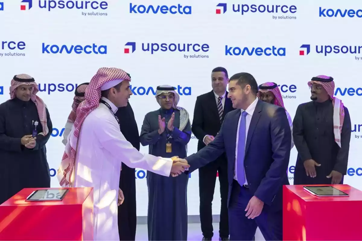 Dos hombres se dan la mano en un evento corporativo, rodeados de otras personas que aplauden, con logotipos de "upsource" y "konvecta" en el fondo.