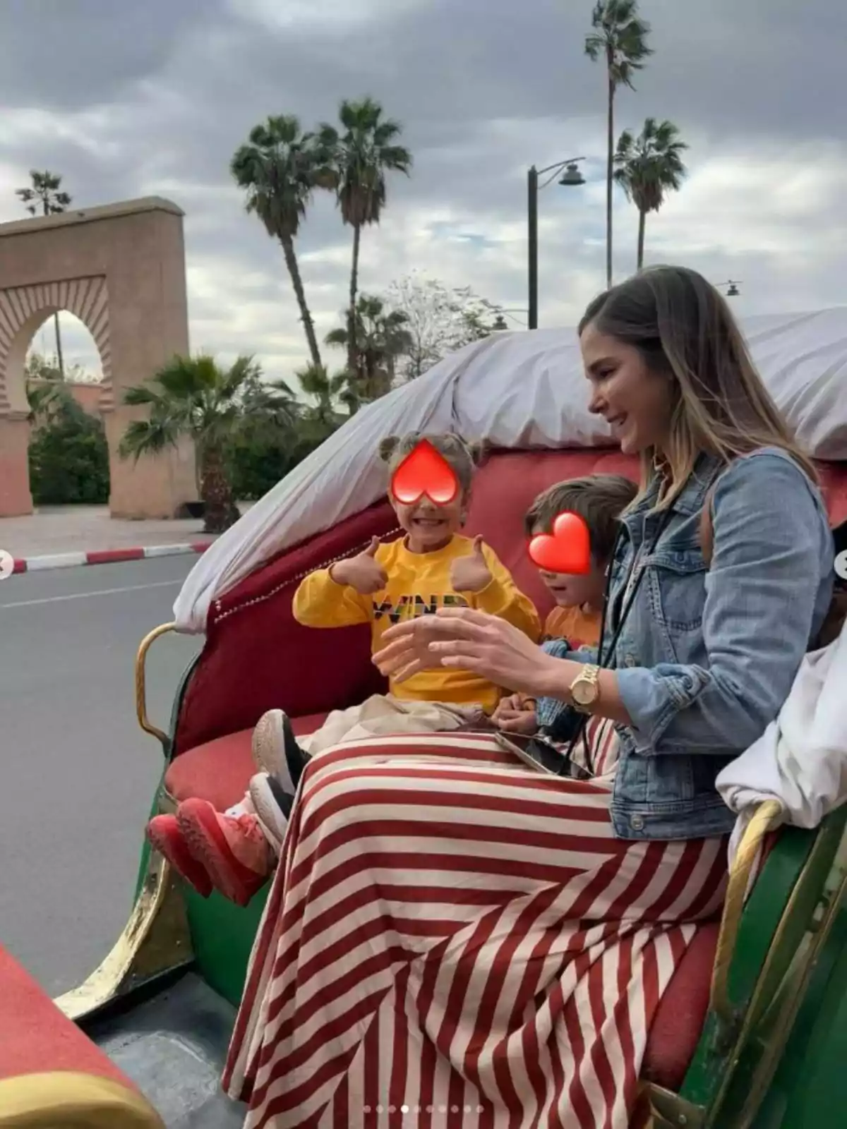 Natalia Sánchez y sus dos hijos, con los rostros cubiertos por emojis de corazón, están sentados en un carruaje al aire libre, con palmeras y un arco de fondo. Post de Natalia Sánchez en Instagram el 31 de marzo de 2024