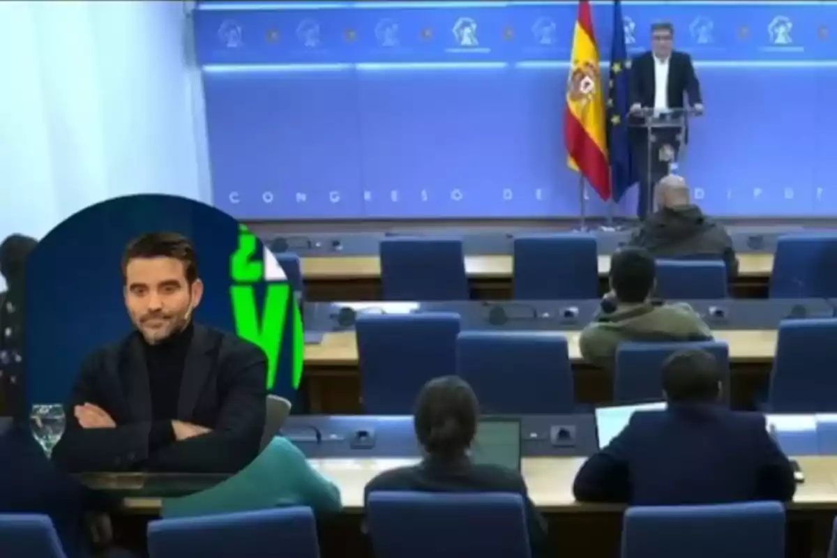 A person at a podium in a conference room with a Spanish flag and a circular screen showing a man with a beard and a black jacket.