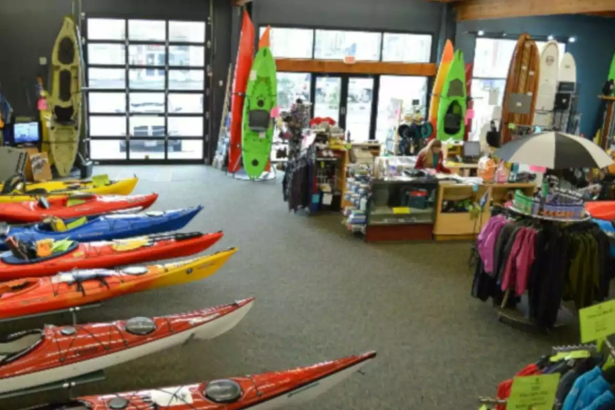 A sporting goods store with brightly colored kayaks lined up on the floor, surfboards and kayaking gear on display, as well as clothing and accessories, while a person attends at the counter. A sporting goods store with brightly colored kayaks lined up on the floor, surfboards and kayaking gear on display, as well as clothing and accessories, while a person attends at the counter.