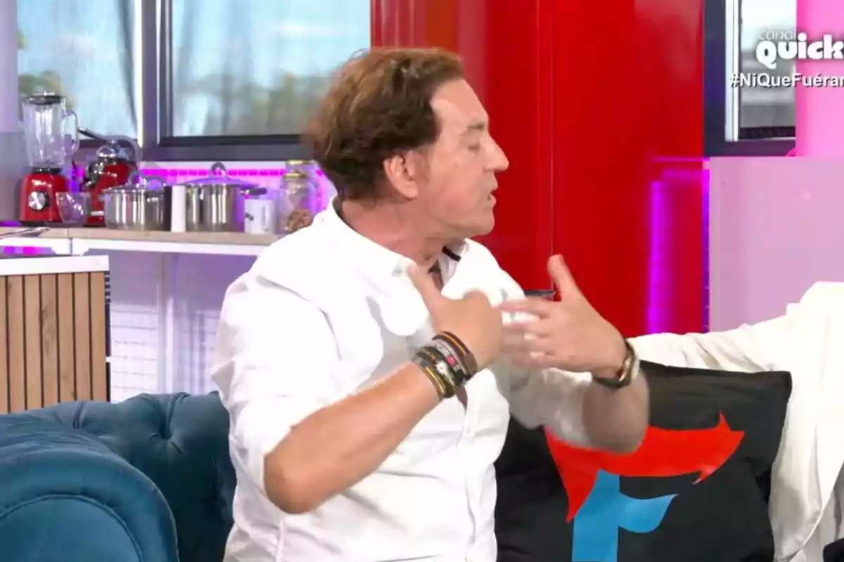 Pipi Estrada con camisa blanca y varias pulseras en la muñeca está gesticulando mientras habla en 'Ni que fuéramos', con una cocina moderna de fondo y un sofá azul en primer plano.