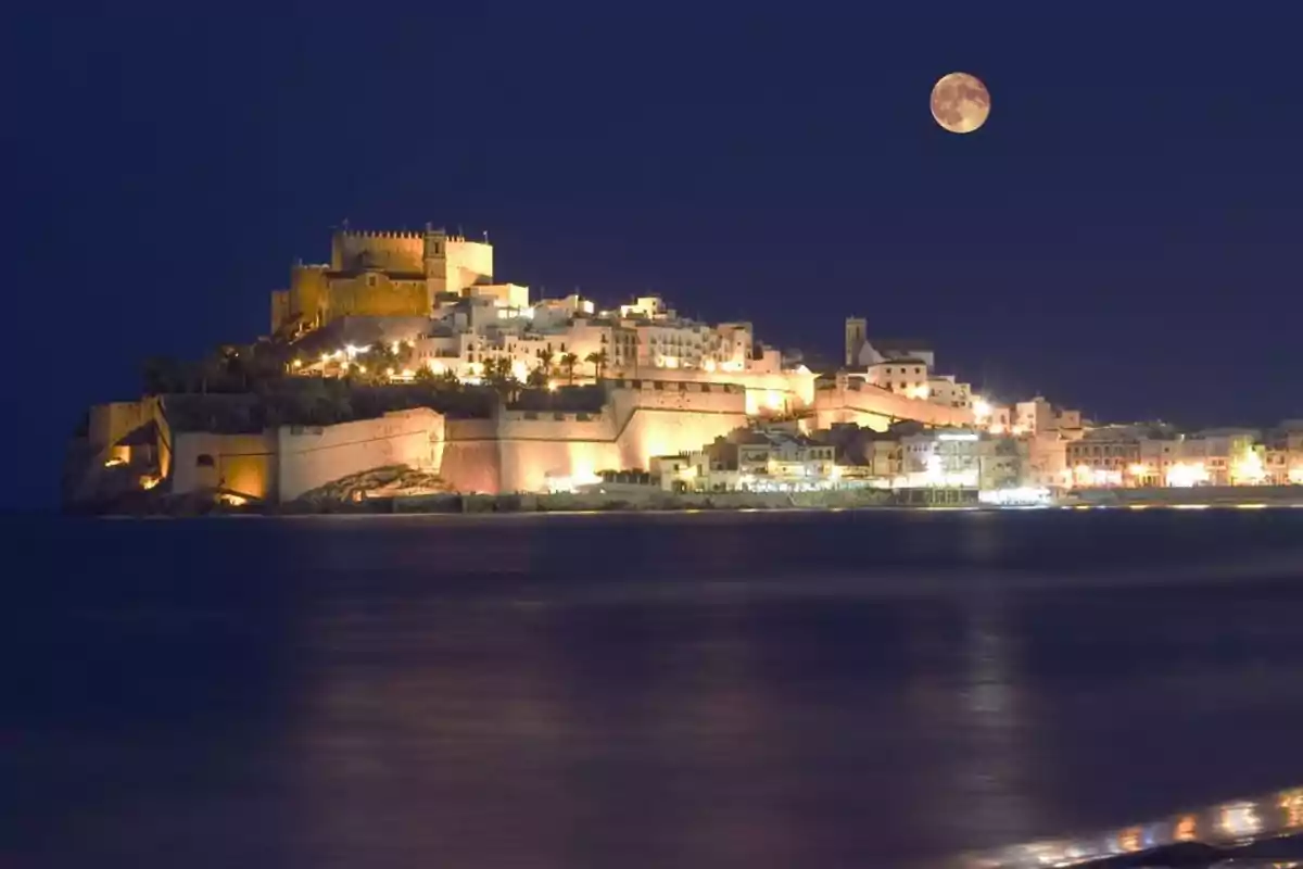 Vista nocturna de un castillo iluminado junto al mar con la luna llena en el cielo.