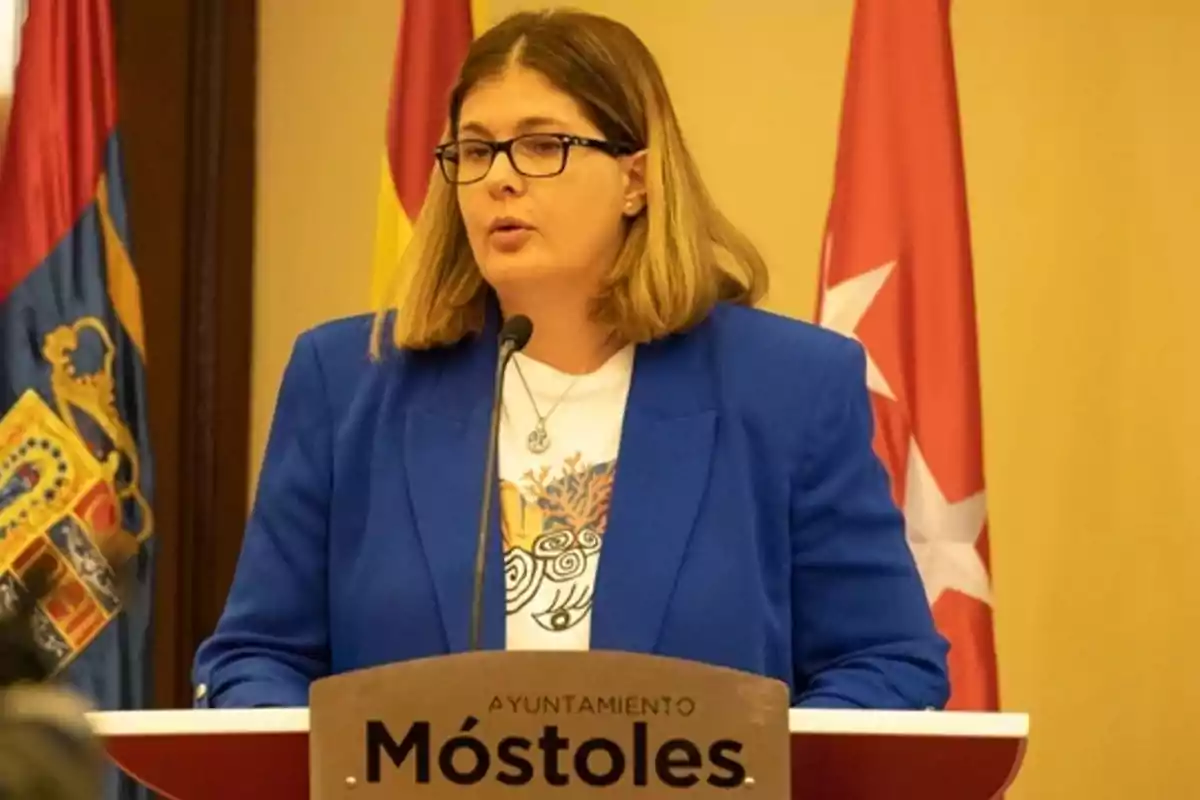 Una persona con gafas y chaqueta azul habla en un podio del Ayuntamiento de Móstoles con banderas de fondo.