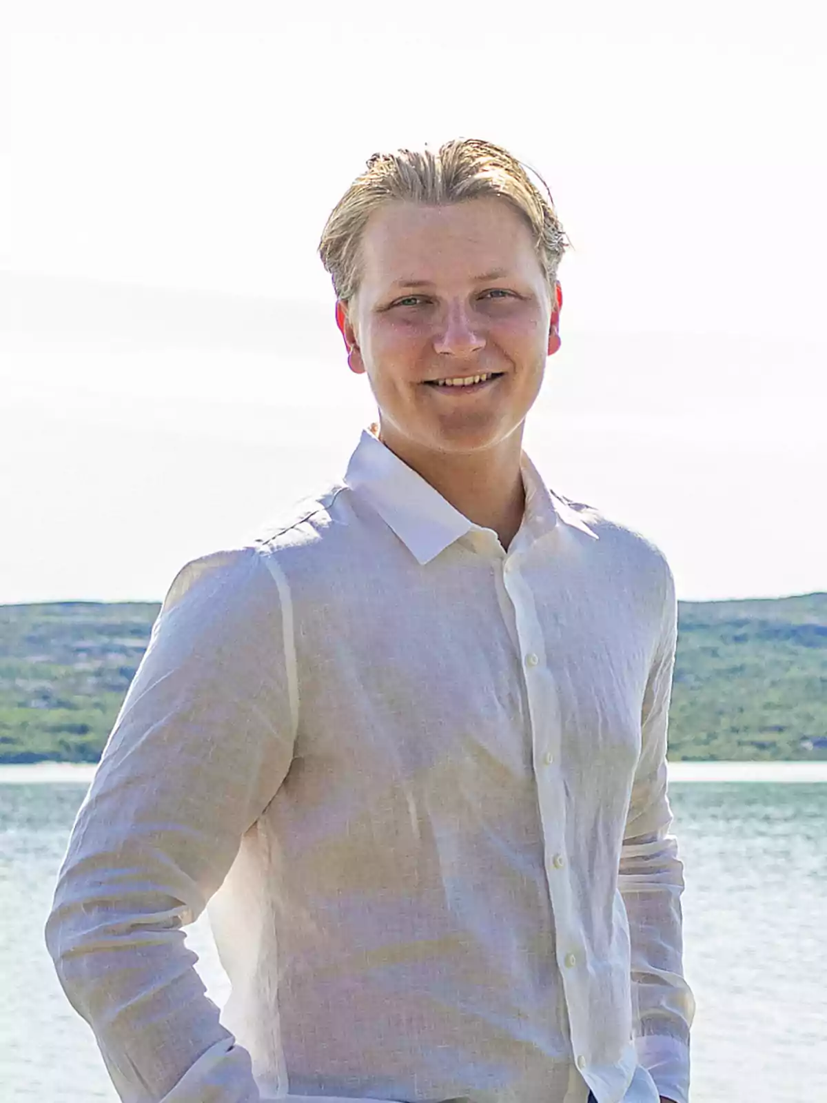 Sverre de Noruega con camisa blanca sonríe frente a un paisaje de agua y colinas. Sverre de Noruega con camisa blanca sonríe frente a un paisaje de agua y colinas.