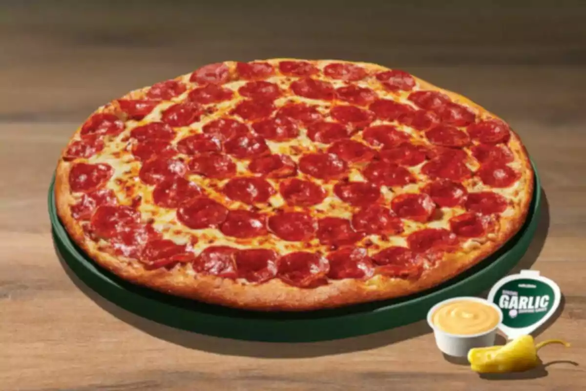Pizza grande de pepperoni sobre una charola verde acompañada de salsas y un chile amarillo sobre una mesa de madera Pizza grande de pepperoni sobre una charola verde acompañada de salsas y un chile amarillo sobre una mesa de madera