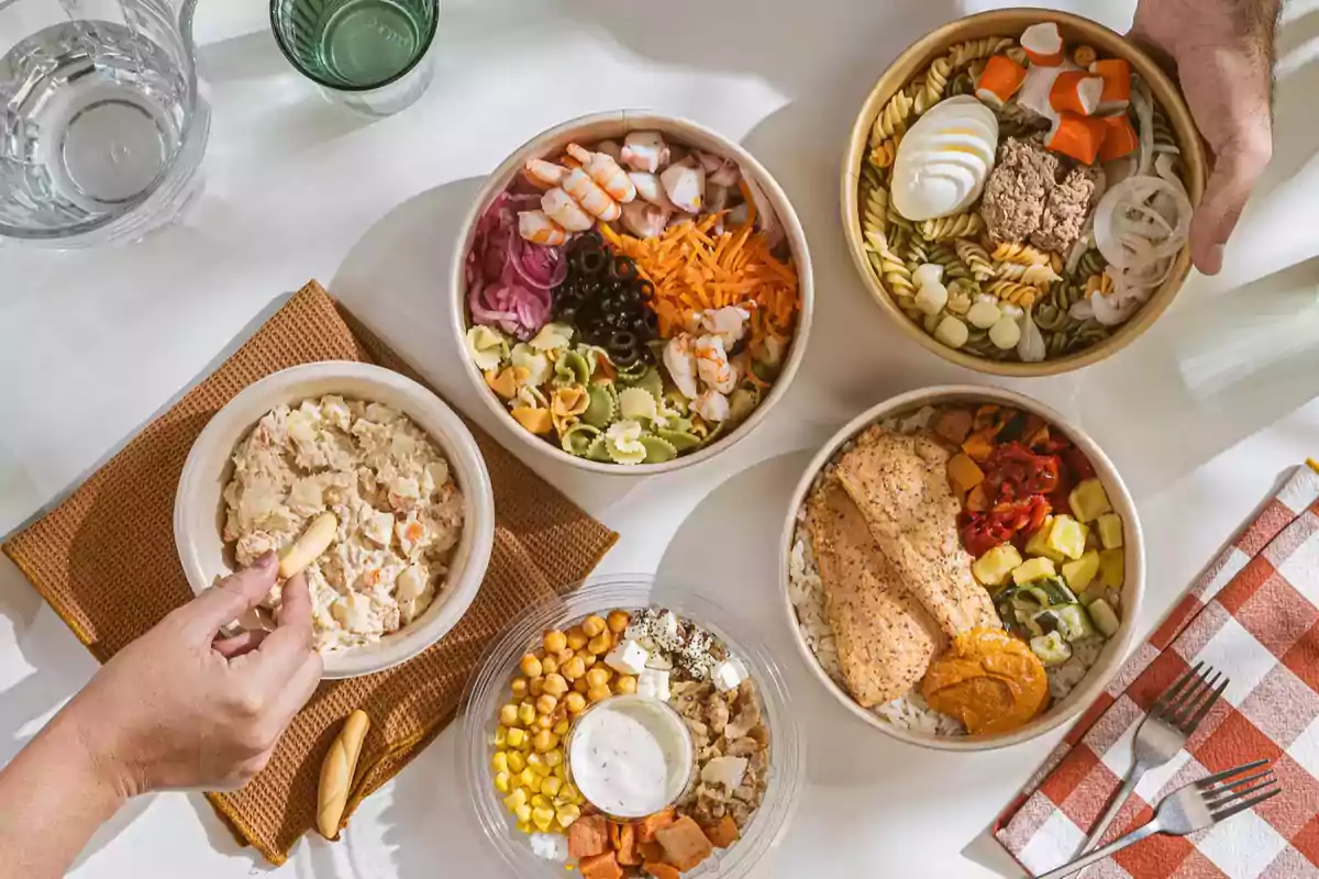 Varias ensaladas y platos de comida en tazones sobre una mesa, con una persona tomando un bocado de ensalada rusa con un palito de pan.