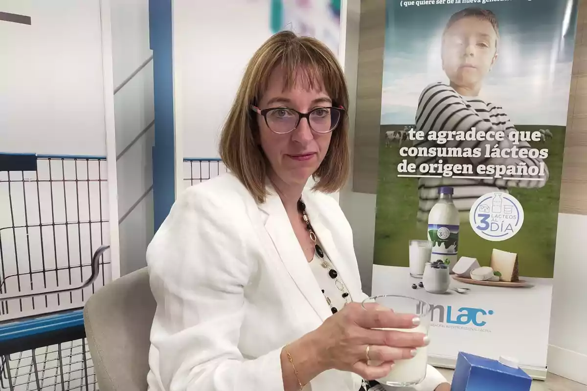 Mujer con gafas y chaqueta blanca sosteniendo un vaso de leche frente a un cartel que promueve el consumo de lácteos de origen español.