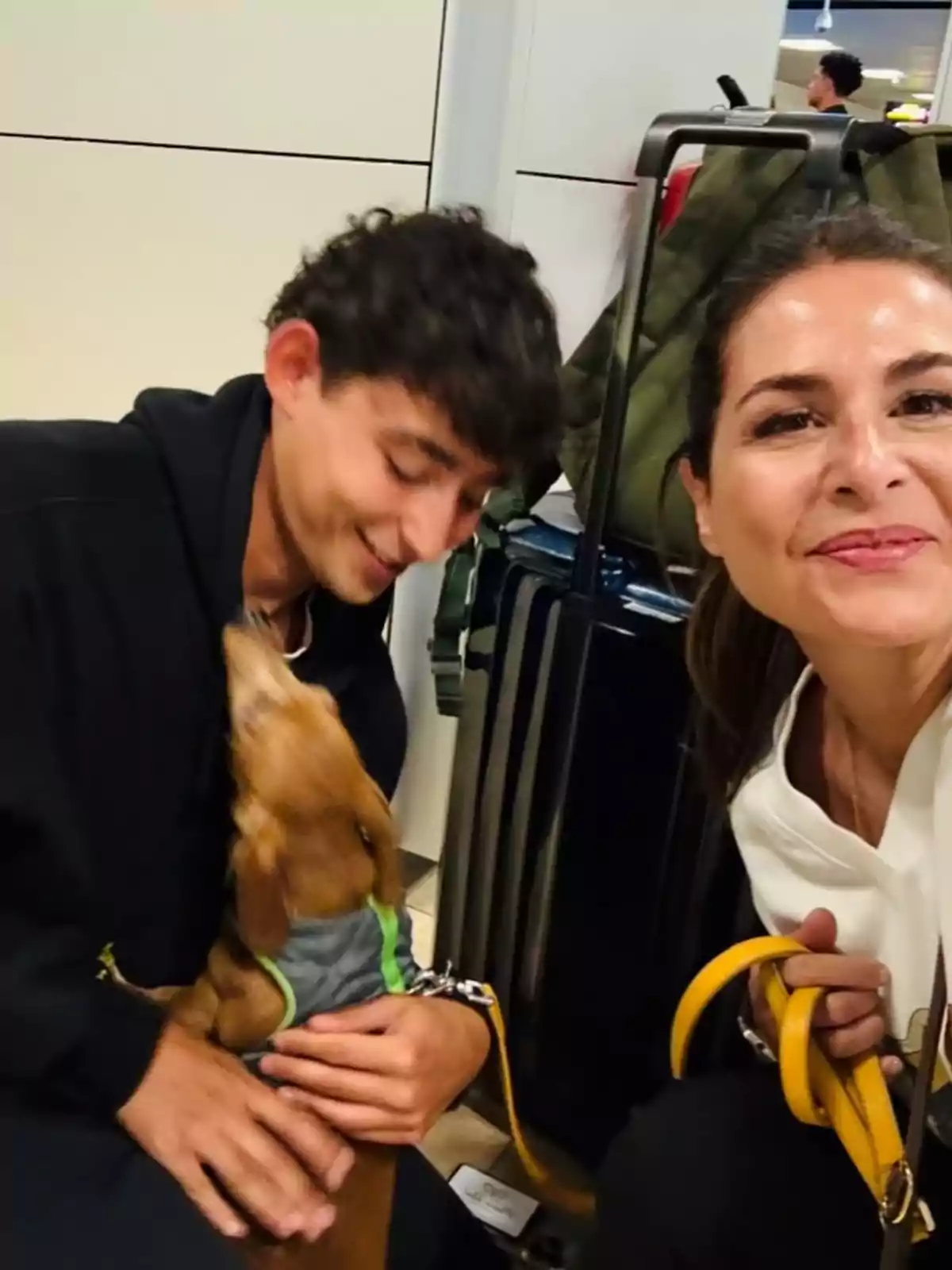 Nuria Roca sostiene una correa amarilla mientras Juan del Val Roca acaricia a un perro pequeño que lleva un chaleco gris, con maletas en el fondo. Nuria Roca sostiene una correa amarilla mientras Juan del Val Roca acaricia a un perro pequeño que lleva un chaleco gris, con maletas en el fondo.