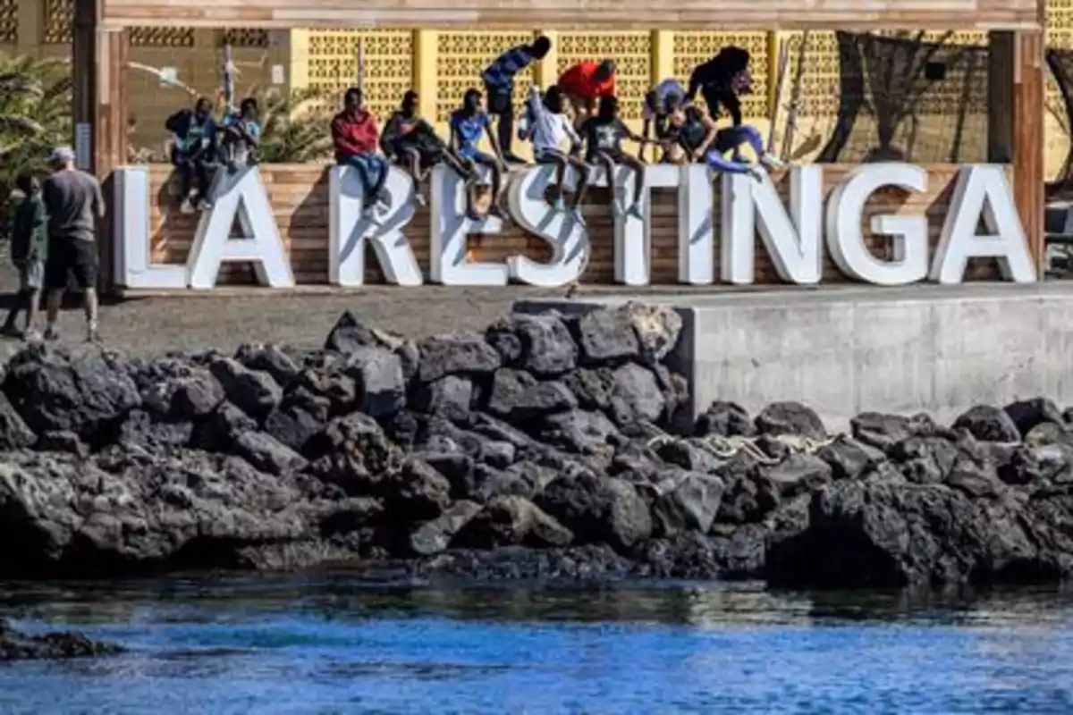 Un grupo de personas está sentado sobre un letrero grande que dice "La Restinga" junto a un muro de rocas y agua.