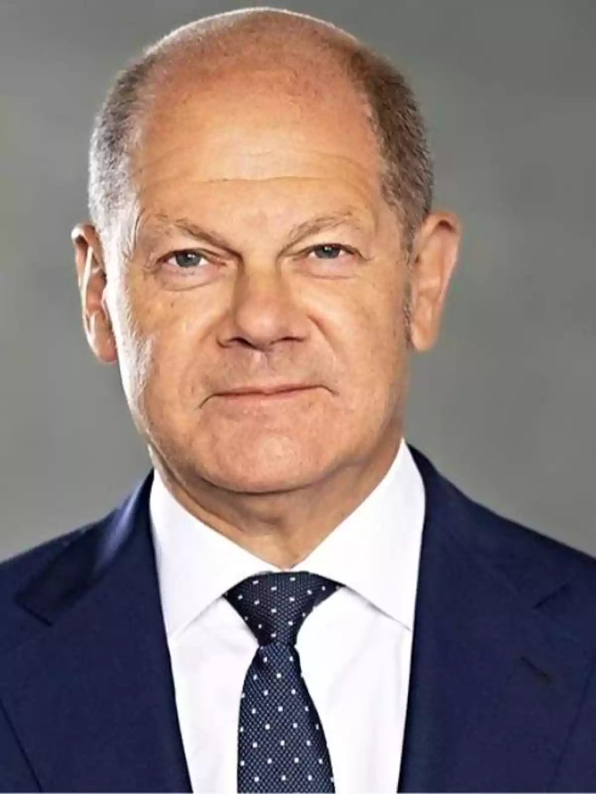 Bald man in a blue suit and polka dot tie against a gray background.